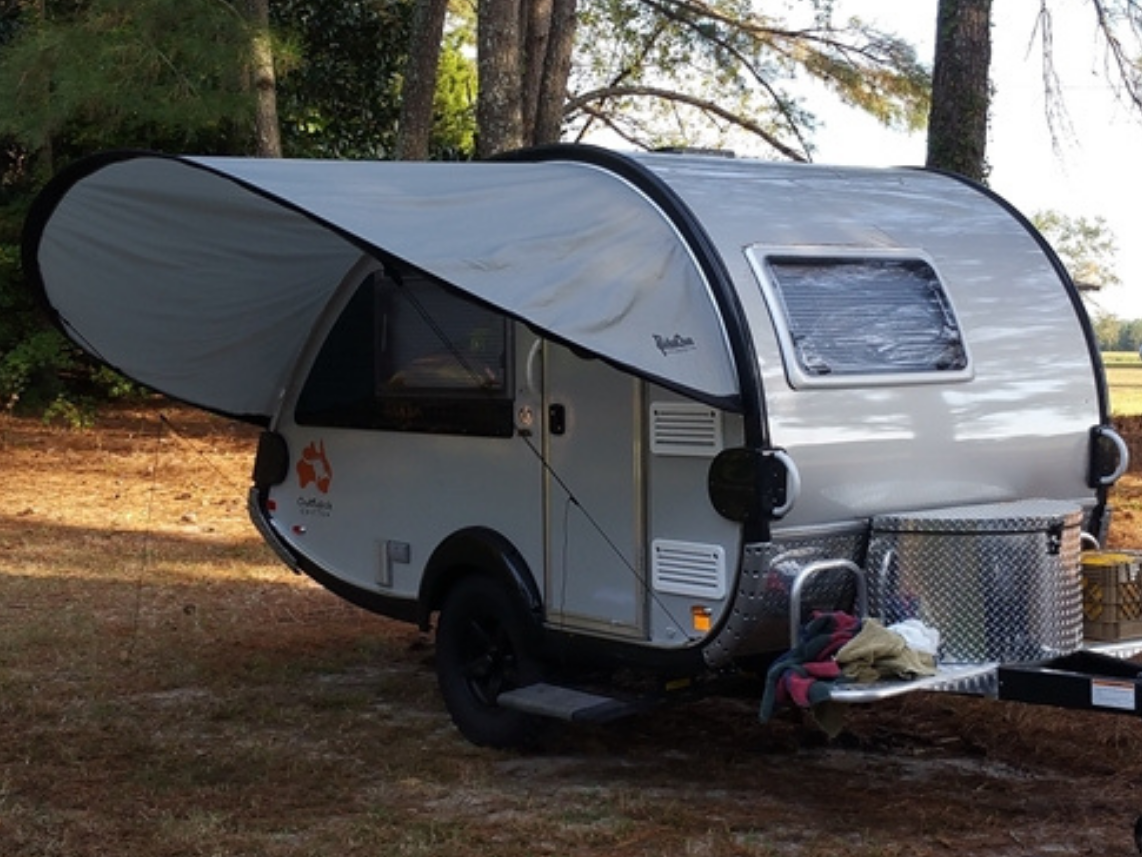 A NuCamp TAB 360 Trailer Visor Accessory by Paha Que Custom provides enhanced outdoor protection for a small teardrop camper, parked under trees on pine needles with camping gear on the metal front platform as sunlight filters through the canopy.