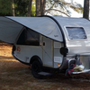 A NuCamp TAB 360 Trailer Visor Accessory by Paha Que Custom provides enhanced outdoor protection for a small teardrop camper, parked under trees on pine needles with camping gear on the metal front platform as sunlight filters through the canopy.