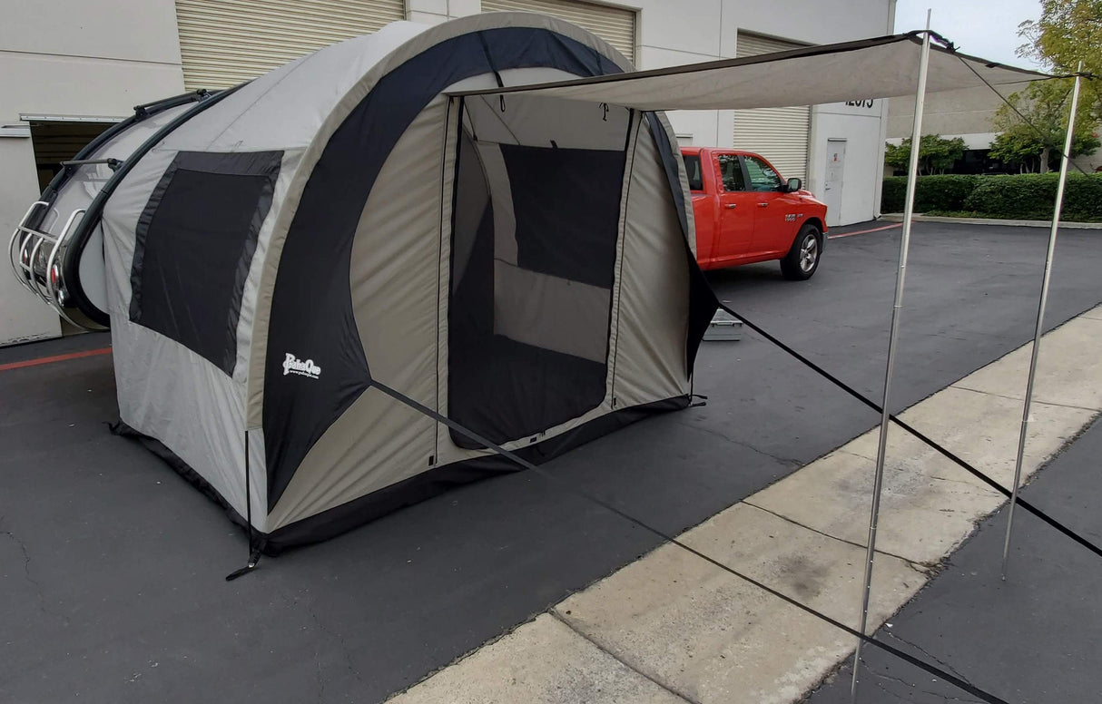 A Paha Que Custom NuCamp TAB 320 Trailer Side Tent for TAB and Boondock Trailers, in gray and black with an extended awning, is set up beside a red pickup truck near beige industrial buildings.