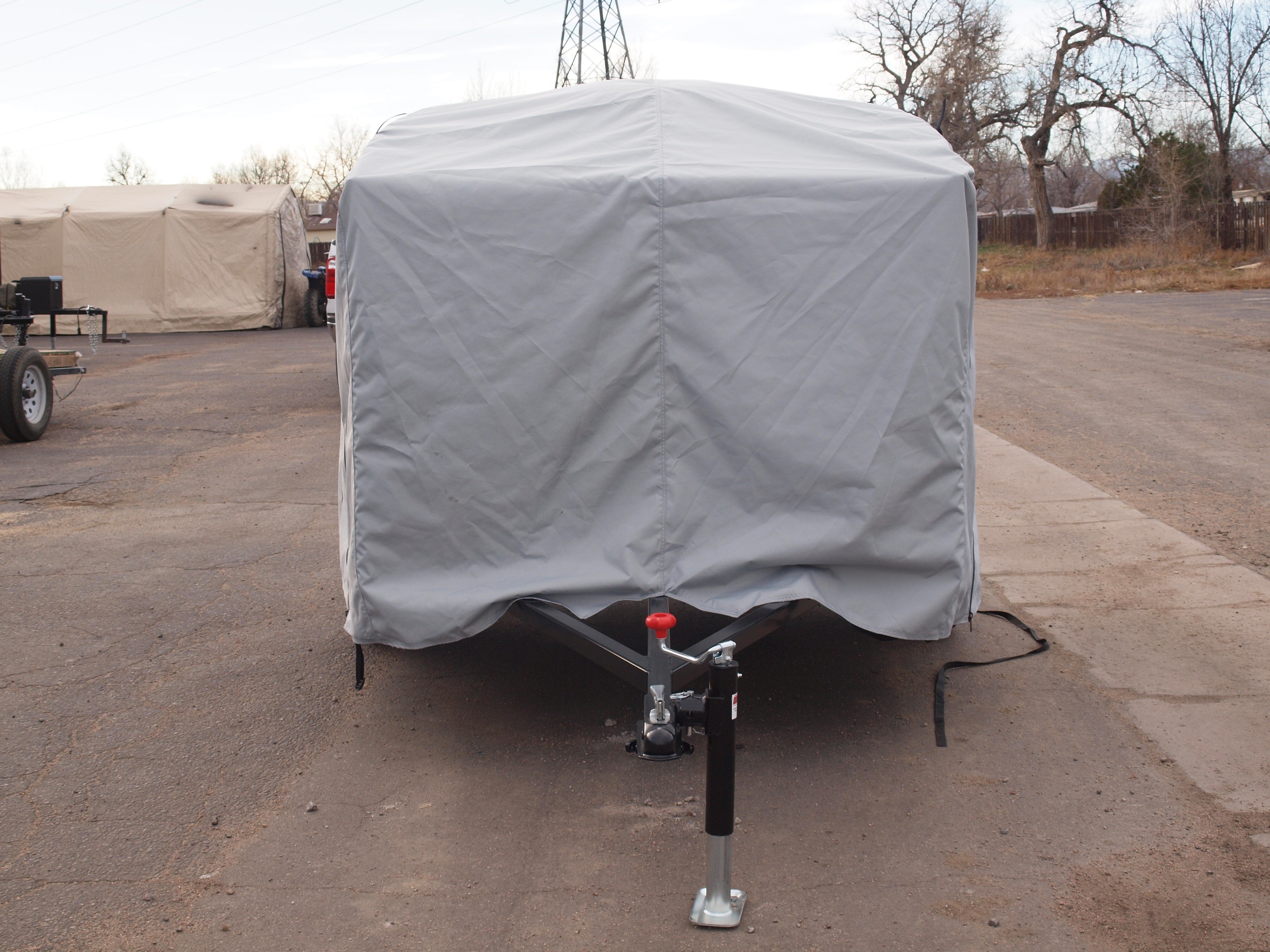 A Hiker Trailer, protected by a Paha Que Custom Hiker Trailer Covers Custom Fit Outdoor Protection, is parked on asphalt. The hitch and jack stand are visible, with other covered vehicles and leafless trees in the background.