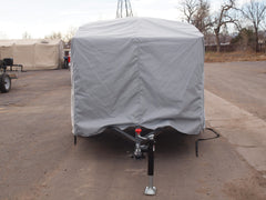 A Hiker Trailer, protected by a Paha Que Custom Hiker Trailer Covers Custom Fit Outdoor Protection, is parked on asphalt. The hitch and jack stand are visible, with other covered vehicles and leafless trees in the background.