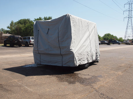 A large vehicle protected by a Paha Que Custom Hiker Trailer Covers Custom Fit Outdoor Protection is parked on an asphalt lot, with other vehicles, trees, and power lines visible in the background under a clear blue sky.