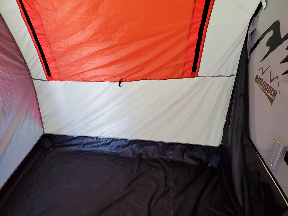 The image shows the inside corner of a Paha Que Custom NuCamp TAB 320 Trailer Side Tent for TAB and Boondock trailers, with a black floor, white walls, an orange upper section, a zippered seam, and part of the BOONDOCK logo on the right.