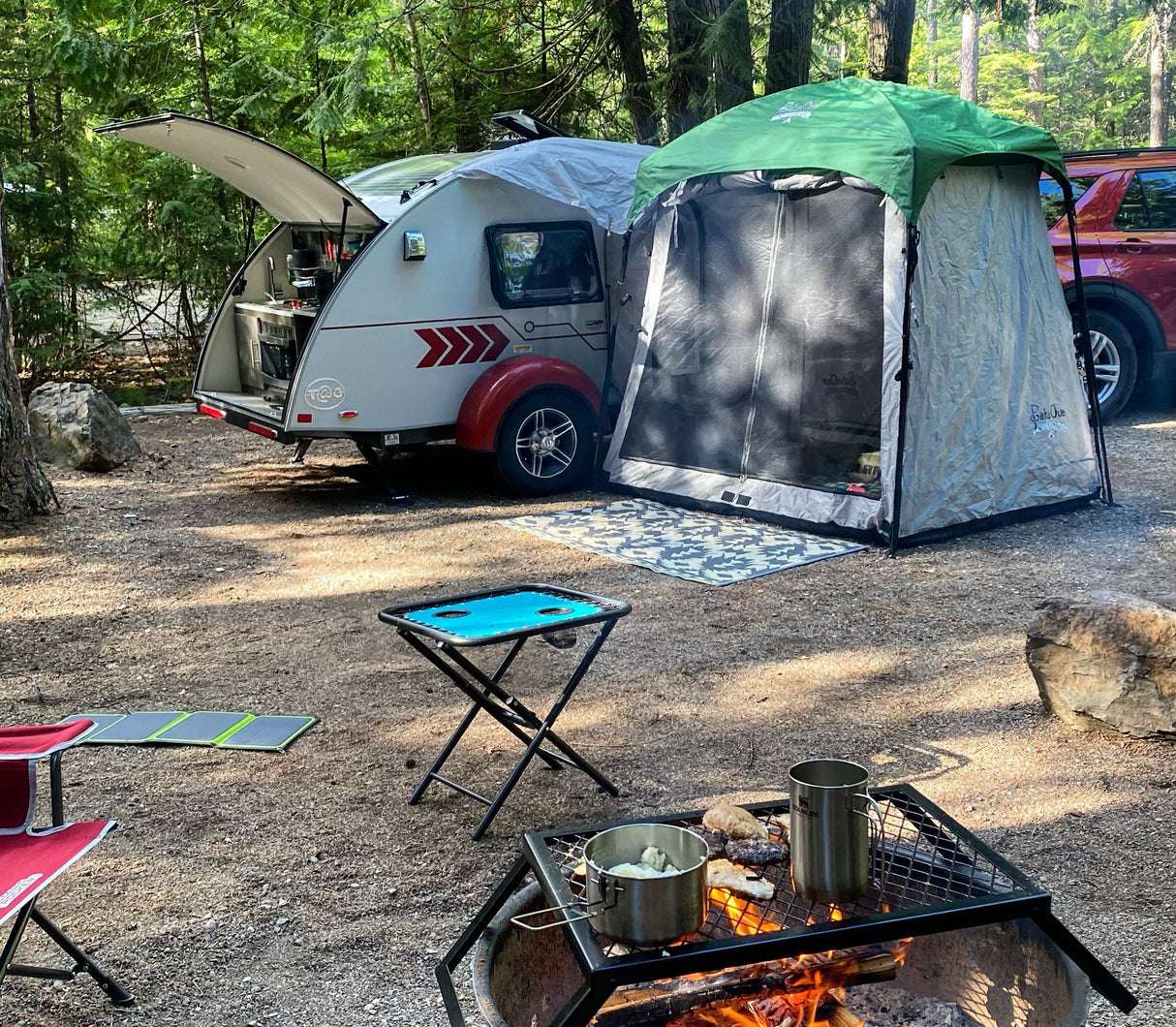 A campsite featuring a Paha Que Custom 5x7 Trailer Side Tent Screen Room, small camper trailer, red car, campfire grilling food, chairs, folding table with camping gear, all surrounded by trees.