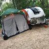 A Paha Que Custom 5x7 Trailer Side Tent Screen Room is set up next to a small white teardrop camper in the woods, with string lights above and camping gear scattered among lush trees at the campsite.