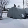 A NuCamp TAG trailer sits in a snowy landscape with snow on the ground and patches on its Paha Que Custom NuCamp TAG Trailer Cover by PahaQue Heavy Duty Weather Proof Protection. Bare trees and a cloudy sky appear in the background.