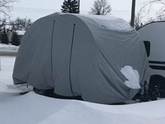 A Paha Que Custom Bushwhacker Plus Trailer Cover (fits models 15DS and 15FK, Made in the USA) protects a trailer as snow rests on top and around it, with snow-covered ground and trees visible under a cloudy sky.