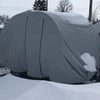 A Paha Que Custom Bushwhacker Plus Trailer Cover (fits models 15DS and 15FK, Made in the USA) protects a trailer as snow rests on top and around it, with snow-covered ground and trees visible under a cloudy sky.