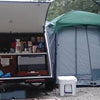 A small camper with its back hatch open shows a compact kitchen setup beside a Paha Que Custom 5x7 Trailer Side Tent Screen Room for Camping and Outdoor Adventures, with camping gear and containers set up on gravel.