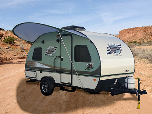 A compact, modern travel trailer equipped with a Paha Que Custom R Pod Trailer Visor is parked on a sandy dirt path in the desert, its extended awning offering shade among rocky hills and scattered vegetation.