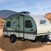 A compact, modern travel trailer equipped with a Paha Que Custom R Pod Trailer Visor is parked on a sandy dirt path in the desert, its extended awning offering shade among rocky hills and scattered vegetation.