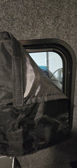 A close-up shows Paha Que Custom Black Out Curtains partially covering a small custom window in a grey, carpeted wall. The black fabric flap with Velcro adds privacy while equipment and a blue item are visible through the window.