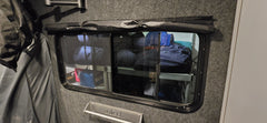 A small window fitted with Paha Que Custom Black Out Curtains reveals organized shelves stocked with camping gear, sleeping bags, and containers inside a compact, gray-carpeted space for privacy.