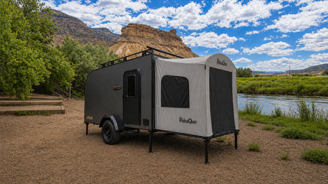 A Toy Hauler Patio Enclosure Ramp Tent by PahaQue Wilderness is attached to a small black and gray camper trailer parked on dirt near a river, surrounded by green trees and rocky hills under a partly cloudy sky.