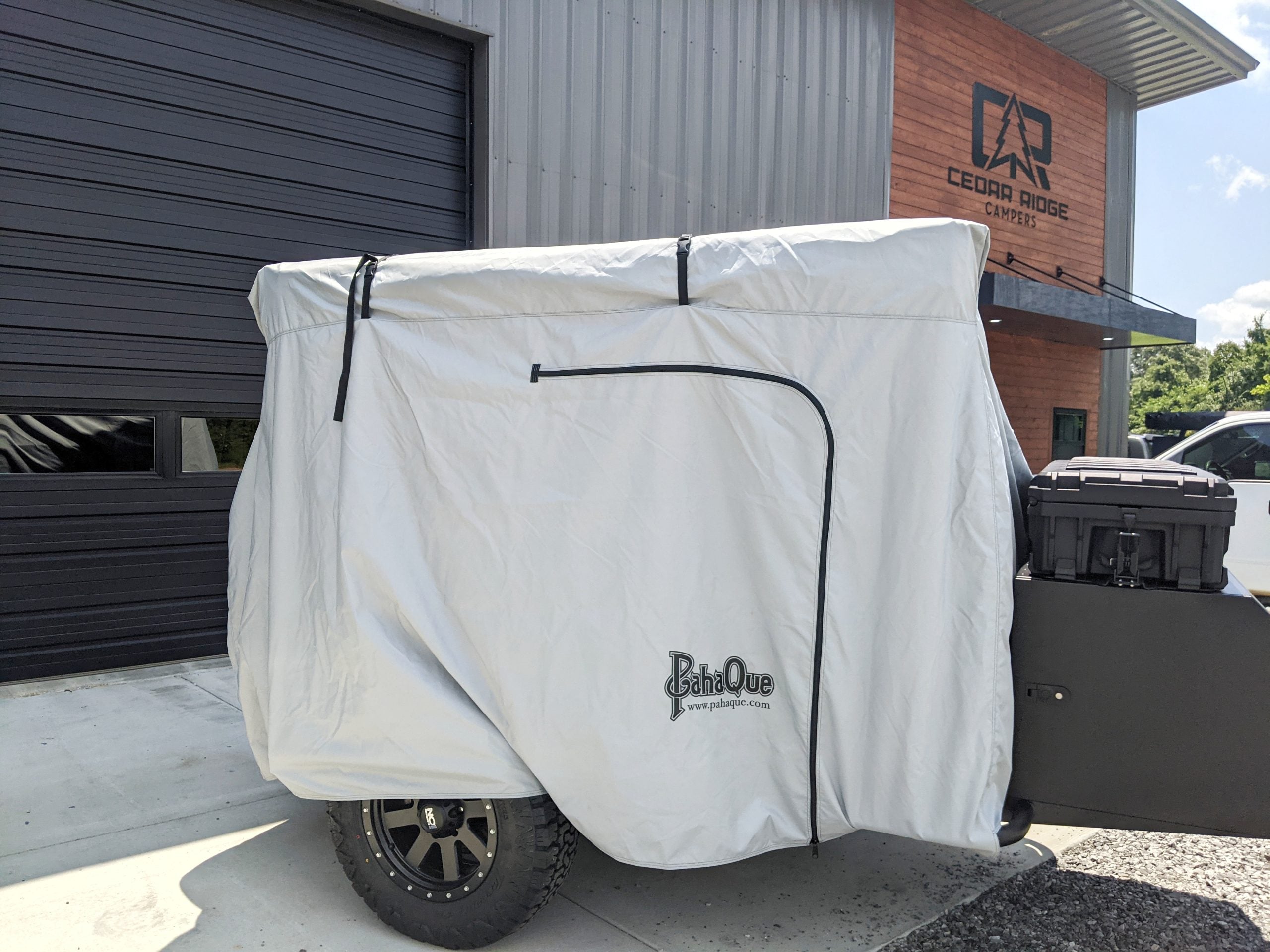 A small trailer with a black wheel and storage box sits outside a building marked Cedar Ridge, protected by the Paha Que Custom Vega XT/AT Cover with Zippered Door Opening.