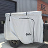 A small trailer with a black wheel and storage box sits outside a building marked Cedar Ridge, protected by the Paha Que Custom Vega XT/AT Cover with Zippered Door Opening.