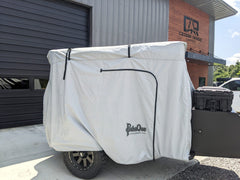 A small trailer with a black wheel and storage box sits outside a building marked Cedar Ridge, protected by the Paha Que Custom Vega XT/AT Cover with Zippered Door Opening.