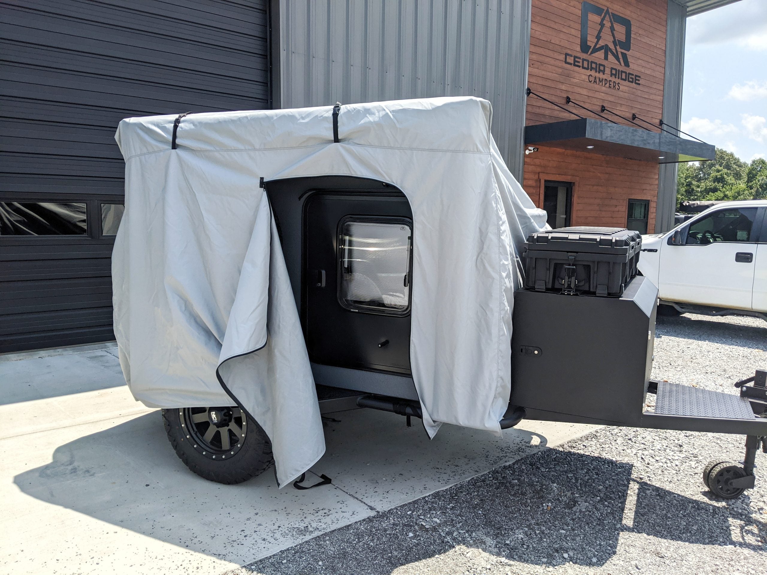 A small black camper trailer is parked outside, partially covered by the PahaQue Vega XT/AT Cover w/ Zippered Door Opening from Paha Que Custom. The light gray cover is pulled back, protecting the trailer while allowing easy access to the window and door.