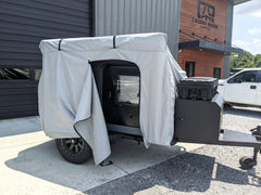 A small black camper trailer is parked outside, partially covered by the PahaQue Vega XT/AT Cover w/ Zippered Door Opening from Paha Que Custom. The light gray cover is pulled back, protecting the trailer while allowing easy access to the window and door.