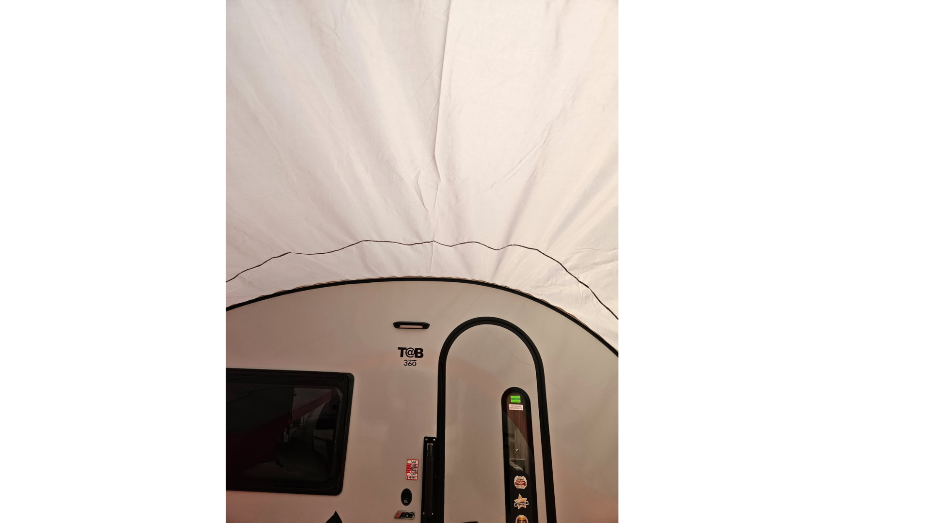 A view from inside a white Paha Que Custom NuCamp TAB 360 Trailer Awning, showing the curved roof, window, and door of a teardrop camper. The awnings waterproof fabric arches above the scene.