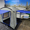 A Paha Que Custom Meerkat Trailer Side Tent for Enhanced Outdoor Adventures and Camping Comfort is set up on a black tarp indoors, next to a white and blue Meerkat trailer with a scenic forest and mountain mural in the background.