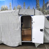 A small teardrop camper sits indoors with its white door open, showing wood flooring inside. A Paha Que Custom Meerkat Trailer Cover Heavy Duty MagnaTuff Fabric is partially removed. A forest and mountain mural is in the background.