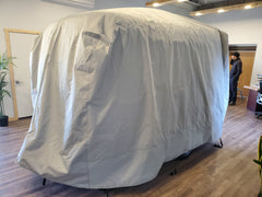 A large object is covered by a gray Paha Que Custom Happier Camper HC1 Cover made from durable MagnaTuff fabric in a room with wooden floors; two people are visible near desks and potted plants in the background.