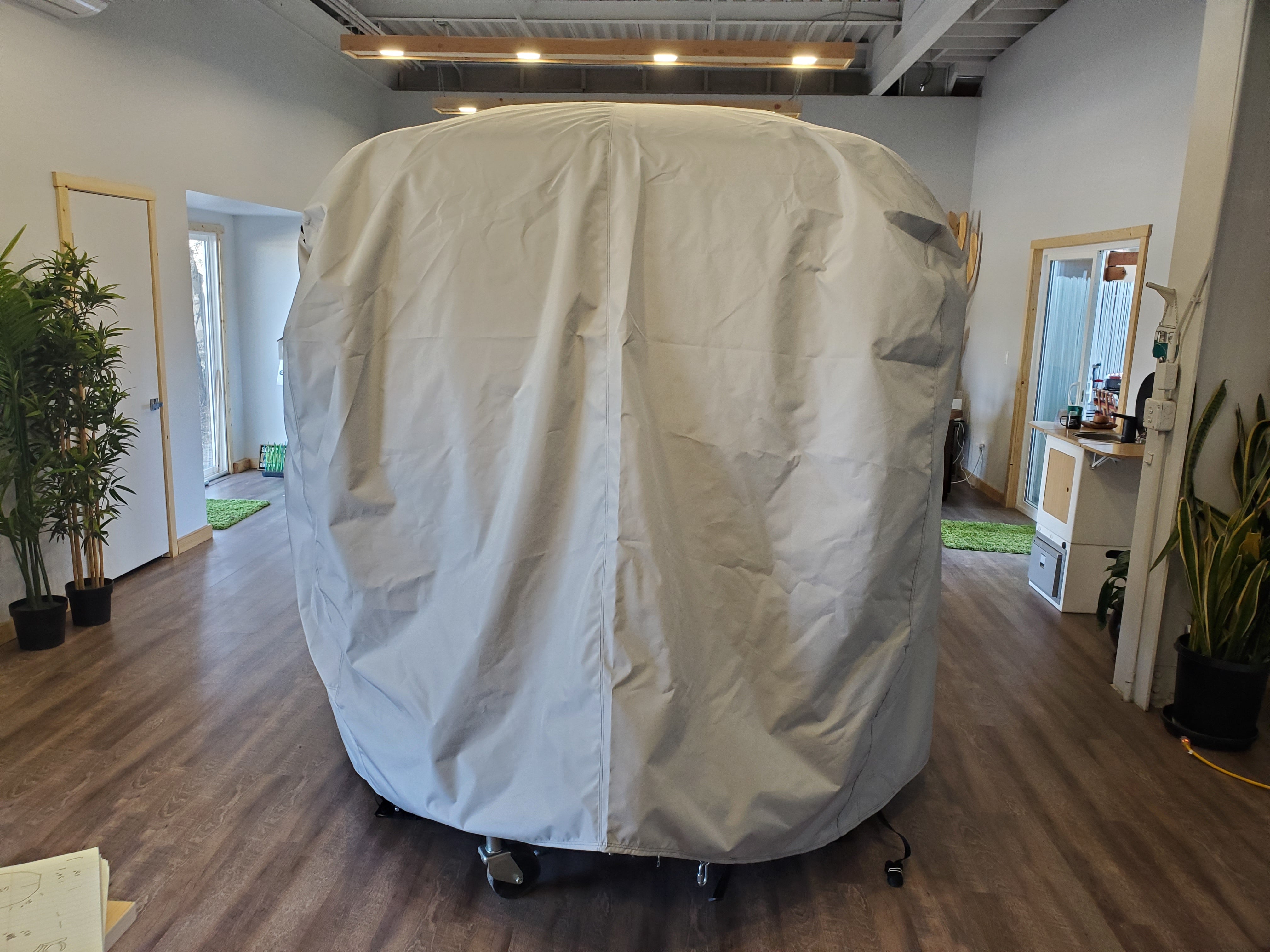 A large gray Paha Que Custom Happier Camper HC1 Cover made of durable MagnaTuff fabric stands on wheels in a modern office with wood floors and plants.