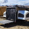 A black camper trailer by Paha Que Custom is set up in a rocky desert, featuring the Toy Hauler Mesh Door/Solid Room Divider for superior insect protection. Camping gear and vehicles are nearby under a partly cloudy sky.