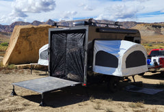 A black camper trailer by Paha Que Custom is set up in a rocky desert, featuring the Toy Hauler Mesh Door/Solid Room Divider for superior insect protection. Camping gear and vehicles are nearby under a partly cloudy sky.