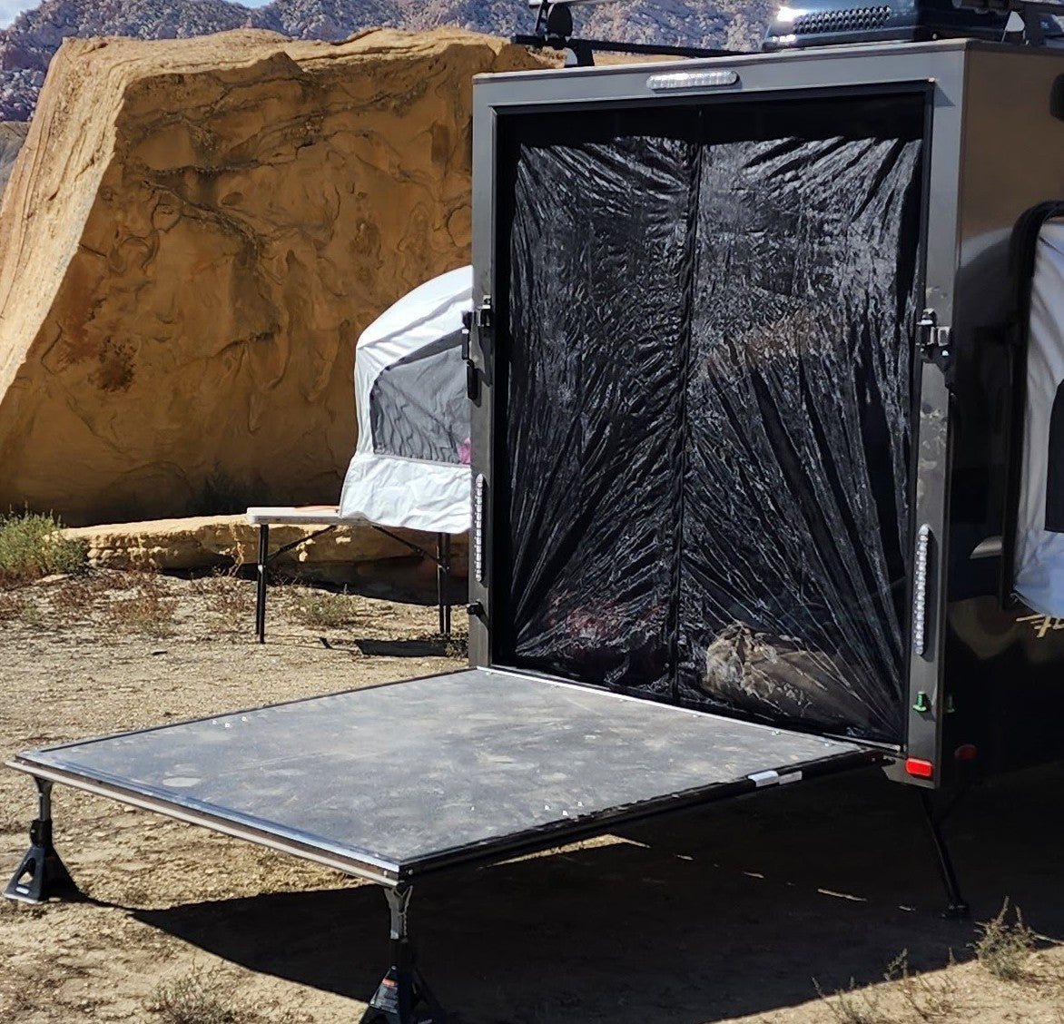 A Paha Que Custom Mesh Door/Solid Room Divider covers the camper trailer's interior for insect protection as its rear hatch opens into a flat platform. Large tan rocks and a white tent dot the surrounding desert landscape.