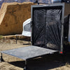 A small camper trailer with its back door open features the Paha Que Custom Toy Hauler Mesh Door/Solid Room Divider for insect protection, forming a ramp with metal legs on sandy ground near a large rock in the desert.
