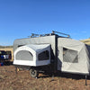 A Paha Que Custom Toy Hauler Patio Ramp Tent, designed for Intech Discover Trailers, is set up on dry, grassy terrain. Nearby are a red pickup truck and various camping gear, with rolling hills and a clear blue sky in the background.