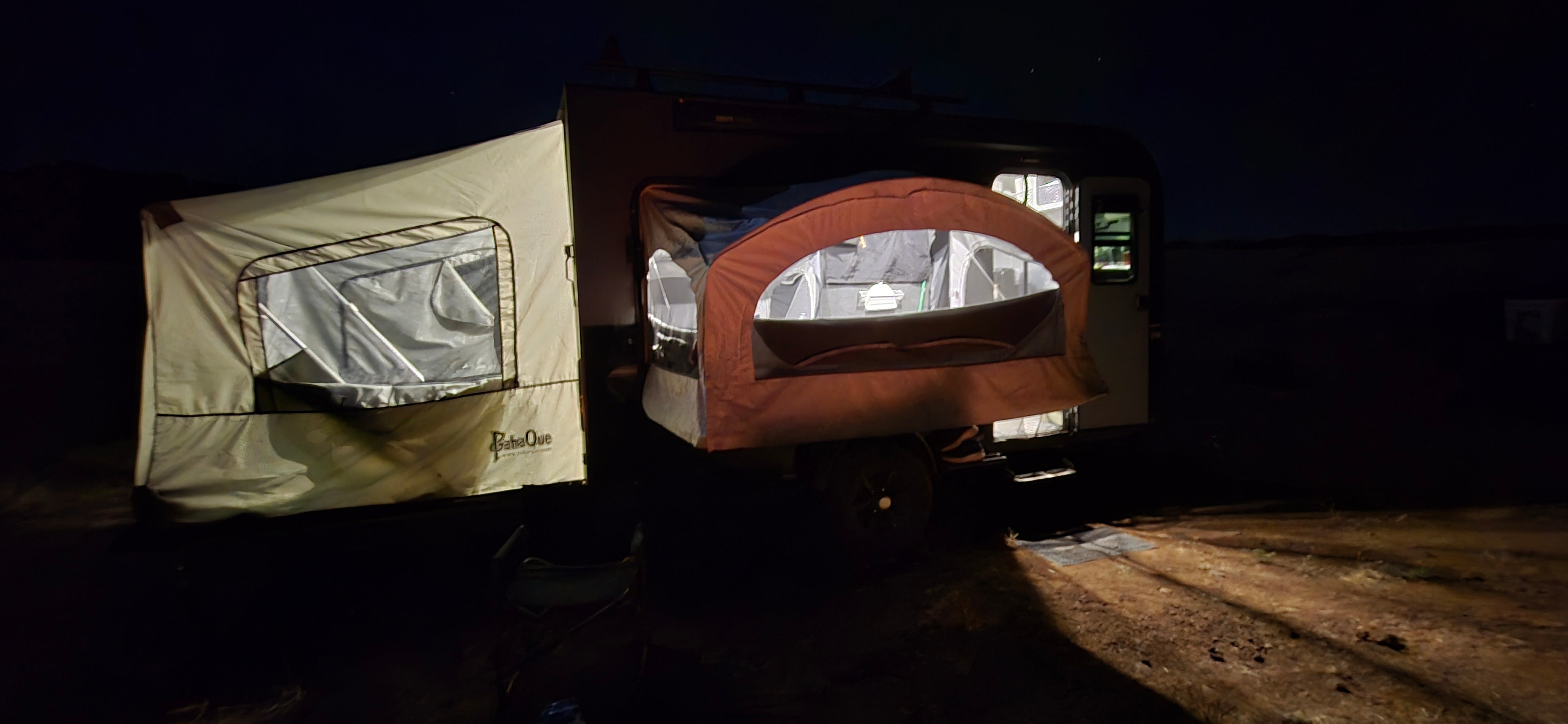 A pop-up camper glows at night with its extended canvas lit from within, while the Paha Que Custom Toy Hauler Patio Ramp Tent by PahaQue for Intech Trailers adds cozy, weatherproof space and enhances the inviting camping atmosphere after dark.