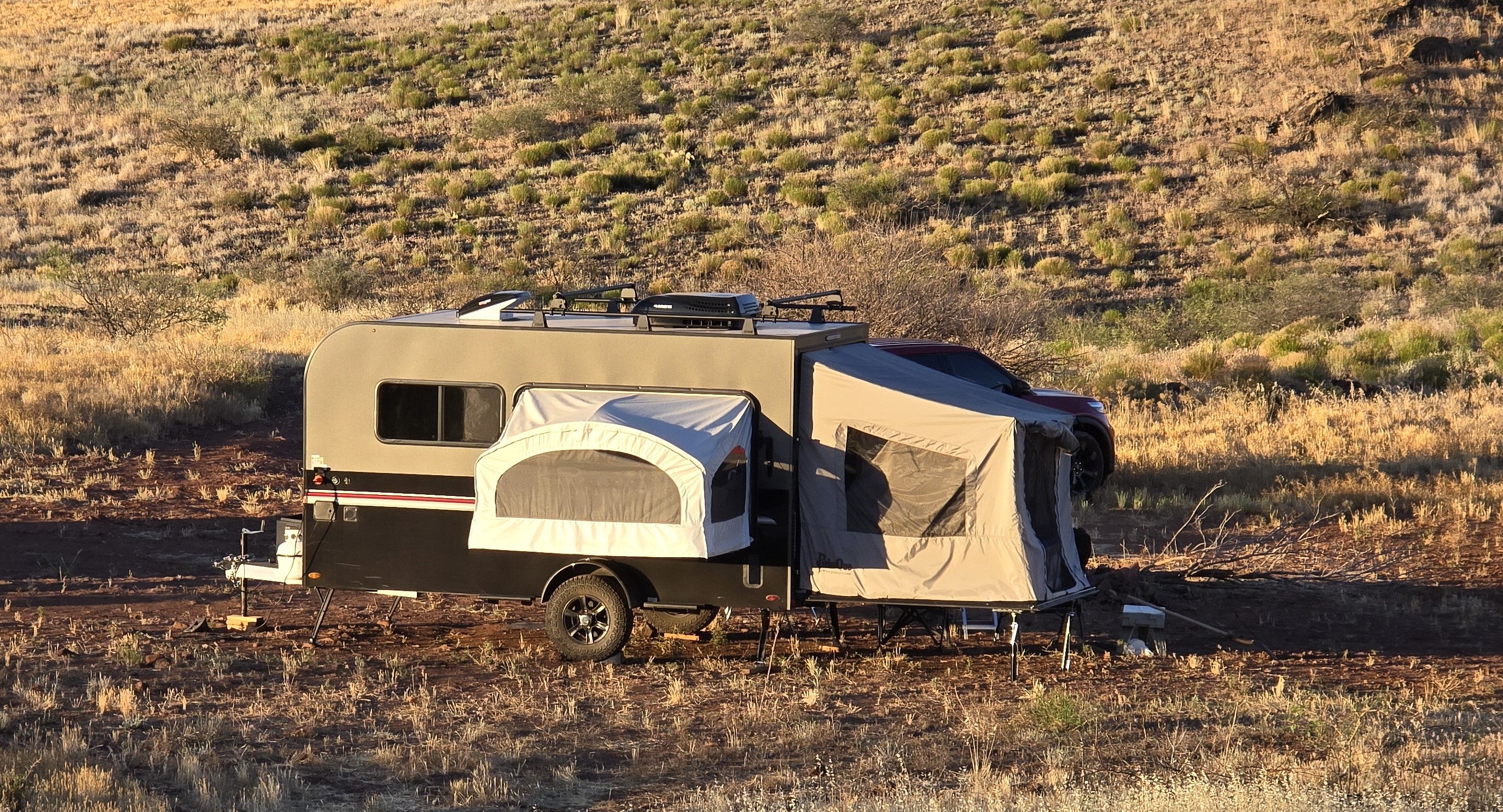 A Toy Hauler Patio Ramp Tent by Paha Que Custom for Intech Trailers is set up on dry grass in a sunny, arid landscape, with a red vehicle partially visible at the front.