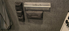 A gray wall with a metal inTech shelf and a PahaQue Wilderness Triple Gear Pouch Heavy Duty Gear Organizer for Trailers attached below, along with two smaller black pouches; part of a black and white object appears on the right.