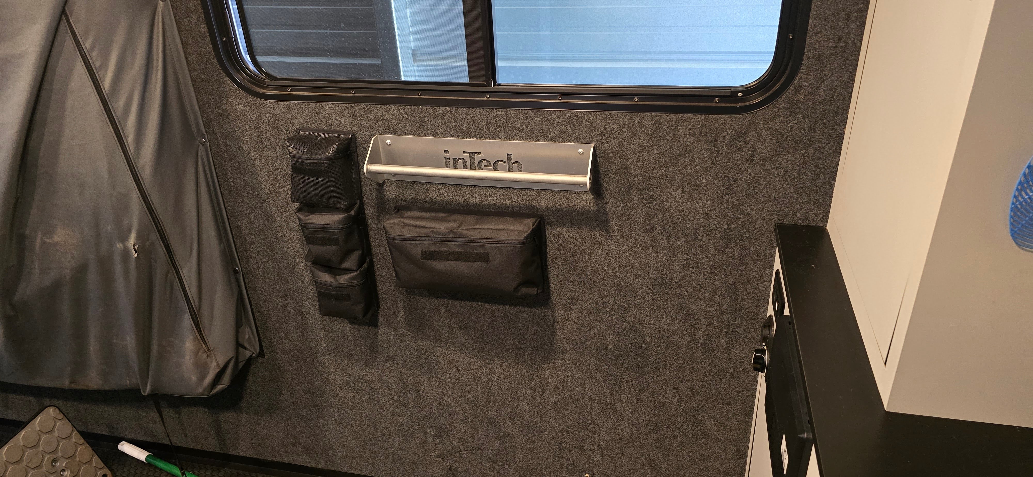 Inside the vehicle, an inTech metal rack holds three PahaQue Wilderness PahaQue Gear Pouches for trailer storage. A window is above, and a whiteboard marker lies on the floor in the lower left corner.