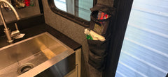 The PahaQue Wilderness Triple Gear Pouch Heavy Duty Gear Organizer for Trailers hangs by a metal sink in a modern RV kitchen, holding toiletries and grooming items. Sunlight from the window highlights this smart black storage solution.