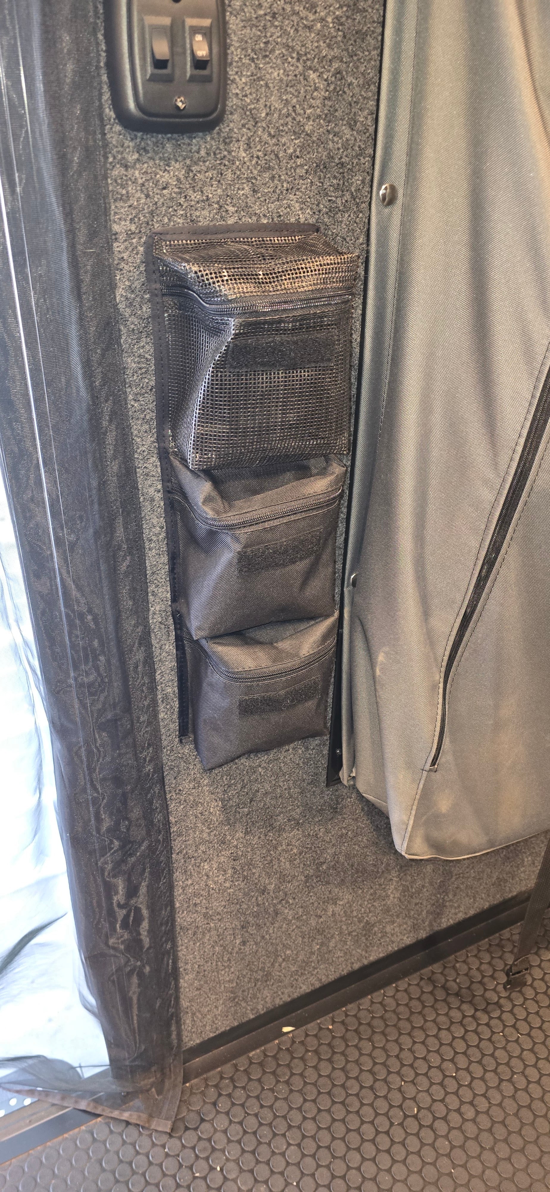 The PahaQue Wilderness Triple Gear Pouch Heavy Duty Gear Organizer is mounted on a gray carpeted wall near a curtain and two electrical switches. Mesh pockets are visible, and the floor has a black hexagonal pattern.