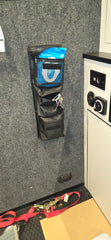 The PahaQue Wilderness Triple Gear Pouch Heavy Duty Gear Organizer for Trailers hangs on a gray wall, holding blue wipes and a key ring. Below is a folded black pouch, with switches on a white cabinet and straps scattered across the black rubber floor.