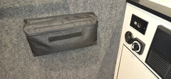 A black PahaQue Gear Pouch by PahaQue Wilderness is mounted on a gray carpeted wall beside a white panel with a power outlet and black control switch, ideal for organizing trailer gear.