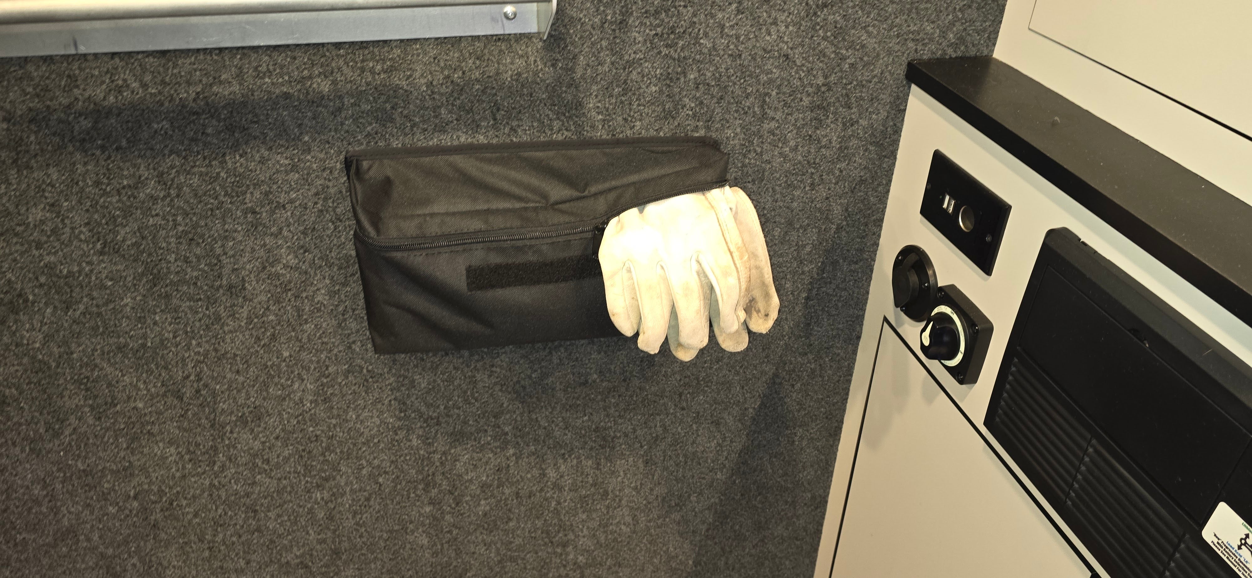 A pair of white work gloves partially stick out of PahaQue Gear Pouches by PahaQue Wilderness, which are mounted on a gray felt wall beside a control panel, offering an efficient trailer gear storage solution.