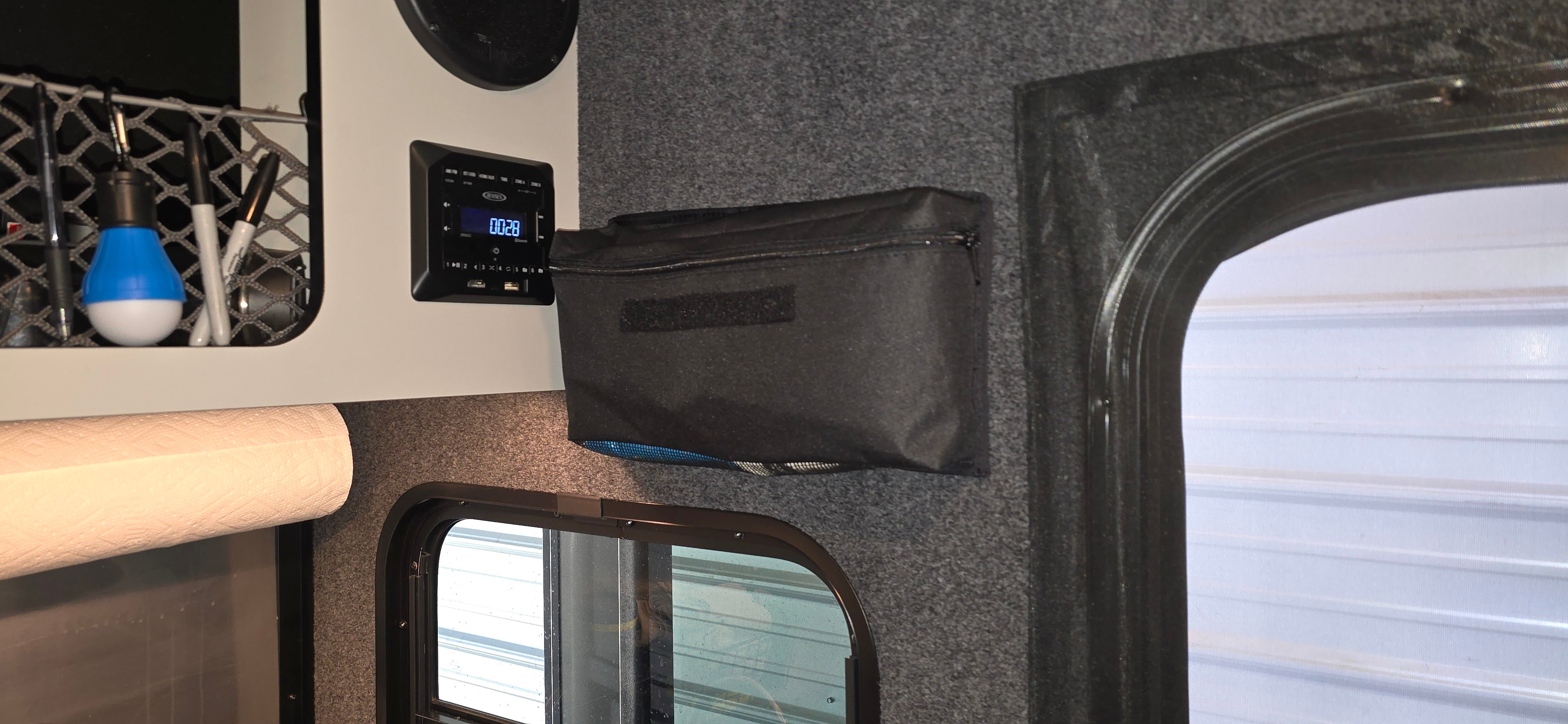 A close-up of a camper interior shows a digital control panel, a PahaQue Gear Pouch by PahaQue Wilderness mounted for trailer gear storage, a window, blue and white bulb, and a paper towel roll on the shelf.