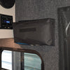 A close-up of a camper interior shows a digital control panel, a PahaQue Gear Pouch by PahaQue Wilderness mounted for trailer gear storage, a window, blue and white bulb, and a paper towel roll on the shelf.