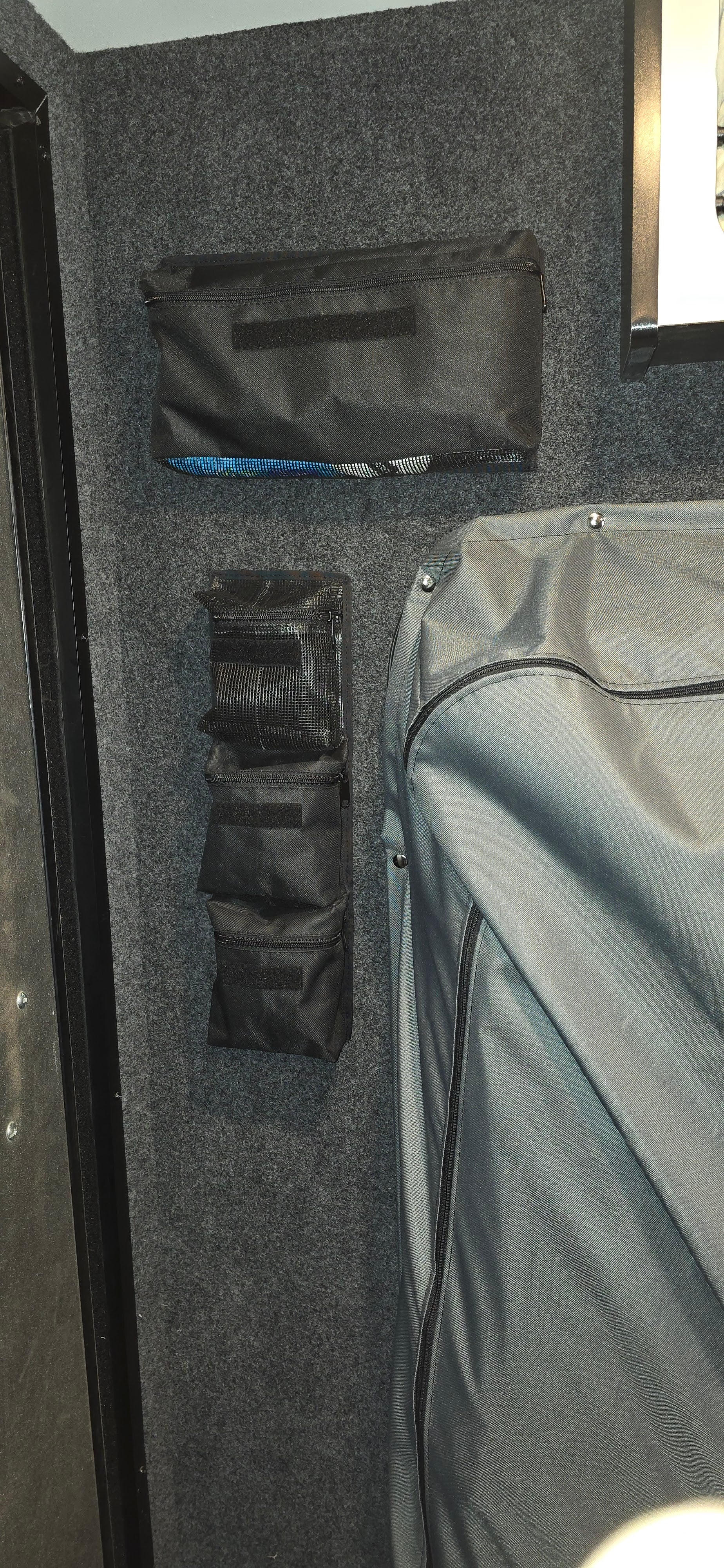 The PahaQue Wilderness PahaQue Gear Pouches, made of black fabric, are mounted on a gray carpeted wall beside a large zippered case—perfect heavy-duty storage for trailer gear in vehicles or equipment rooms.