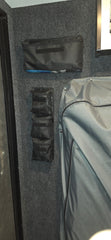 The PahaQue Wilderness PahaQue Gear Pouches, made of black fabric, are mounted on a gray carpeted wall beside a large zippered case—perfect heavy-duty storage for trailer gear in vehicles or equipment rooms.