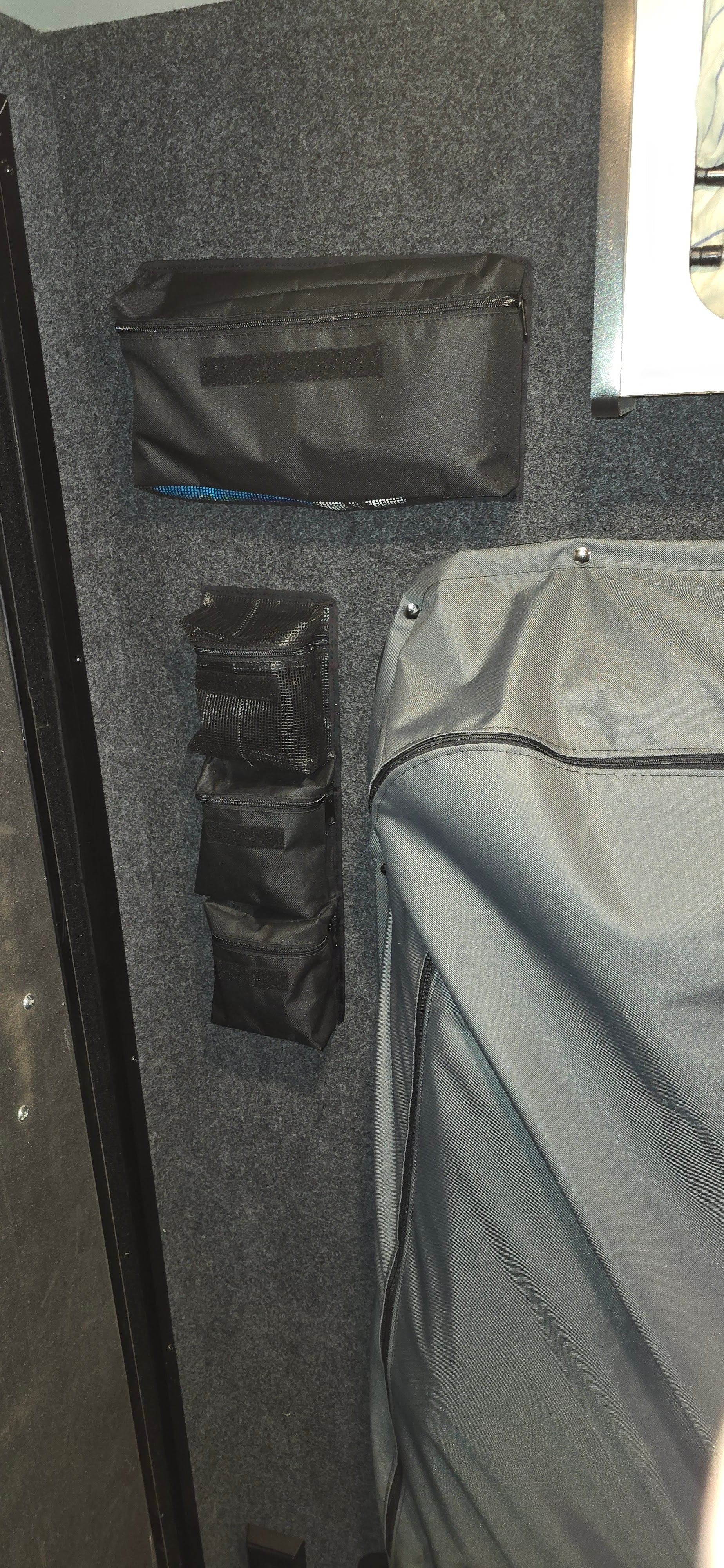 Triple Gear Pouch Heavy Duty Gear Organizer for Trailers by PahaQue Wilderness attached to a dark carpeted wall, with a grey fabric-covered item on the right and part of a framed object visible in the top right corner.