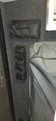 Triple Gear Pouch Heavy Duty Gear Organizer for Trailers by PahaQue Wilderness attached to a dark carpeted wall, with a grey fabric-covered item on the right and part of a framed object visible in the top right corner.