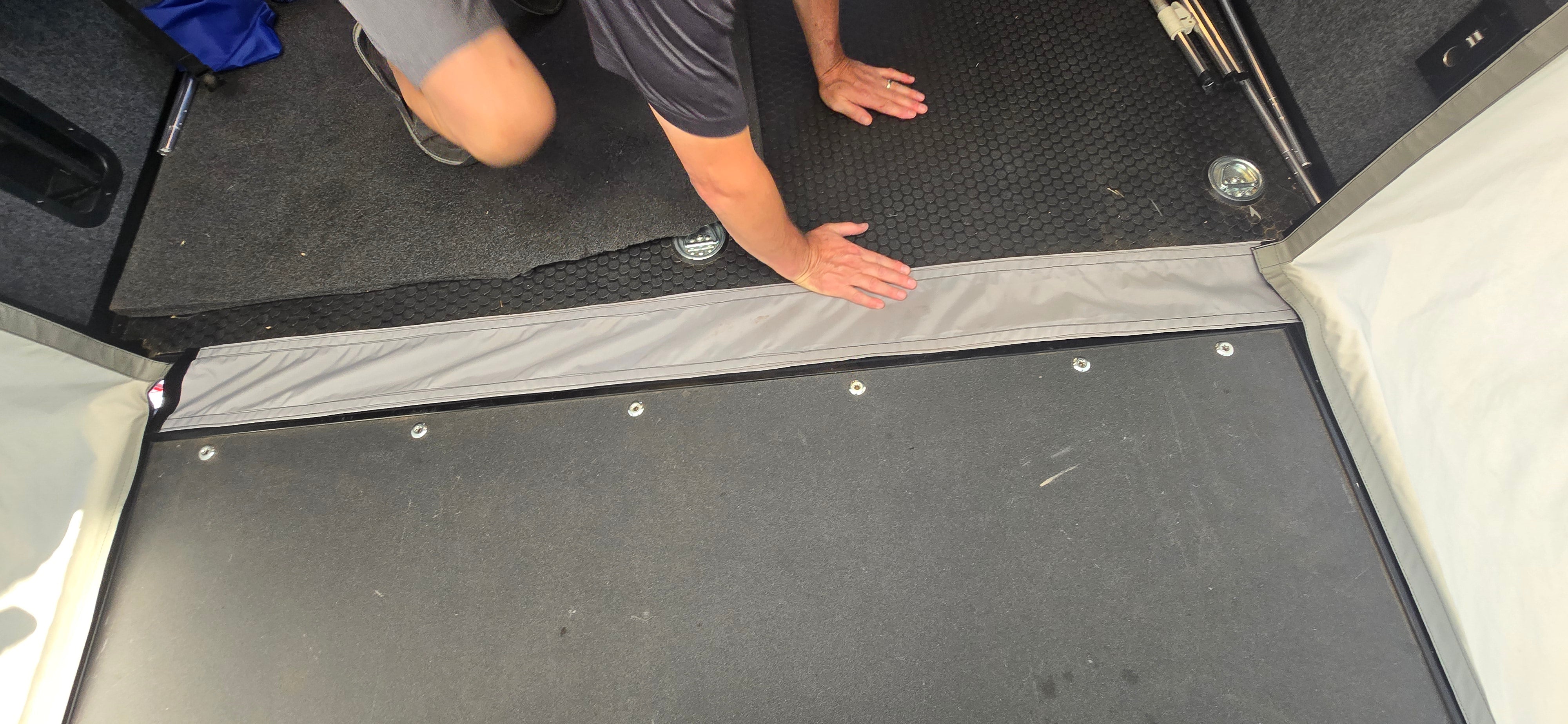A person kneels on a black rubber floor inside an Intech trailer, pressing a fabric seam of the Toy Hauler Patio Ramp Tent by Paha Que Custom. Their face is not visible.