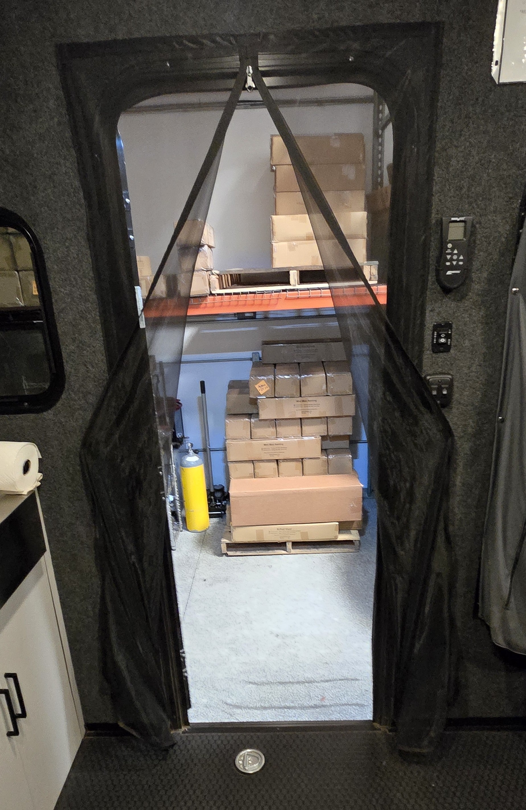 A Paha Que Custom Toy Hauler Mesh Door/Solid Room Divider offers insect protection, revealing storage shelves with boxes inside, while a yellow tank, cleaning tools, dark walls, and a cabinet complete the scene.